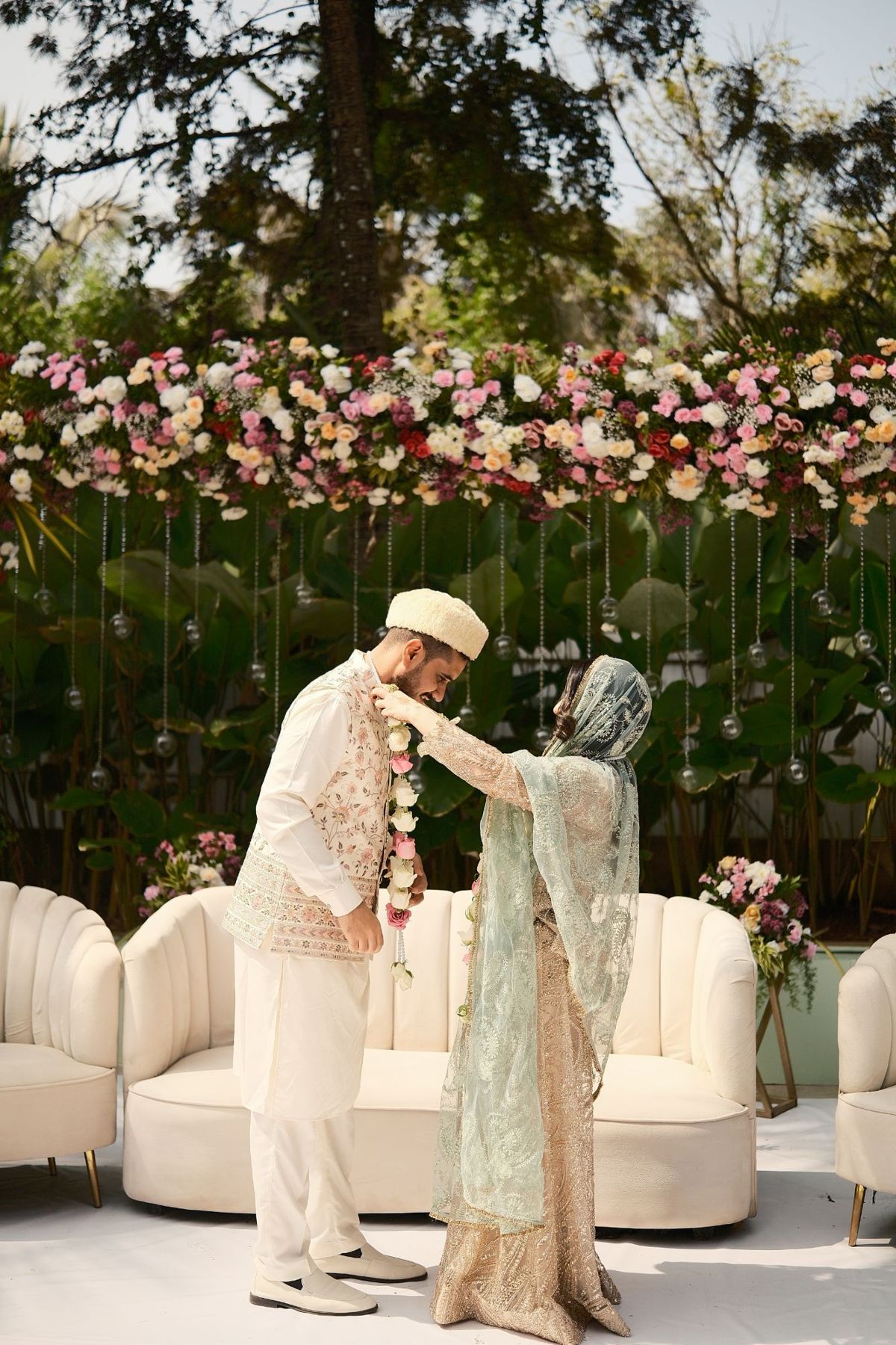 A bride placing a garland on her groom, under the trees